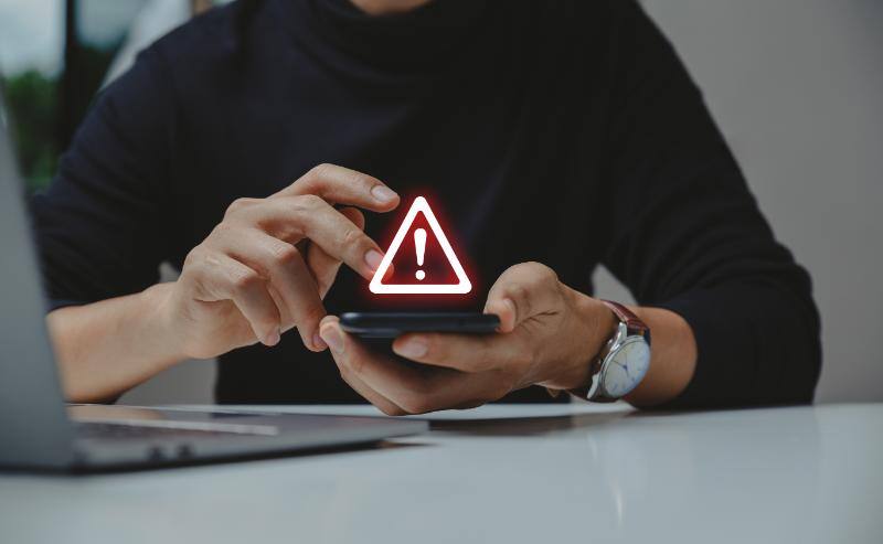 両手で抱えられたスマートフォンに浮かぶ注意マーク